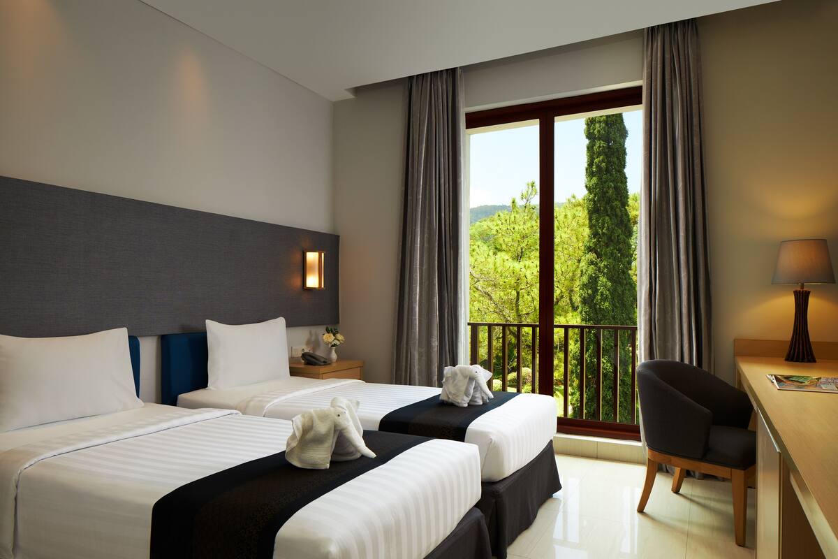 A hotel room with two beds, a desk, and a chair. There is a window with curtains and a view of greenery outside.
