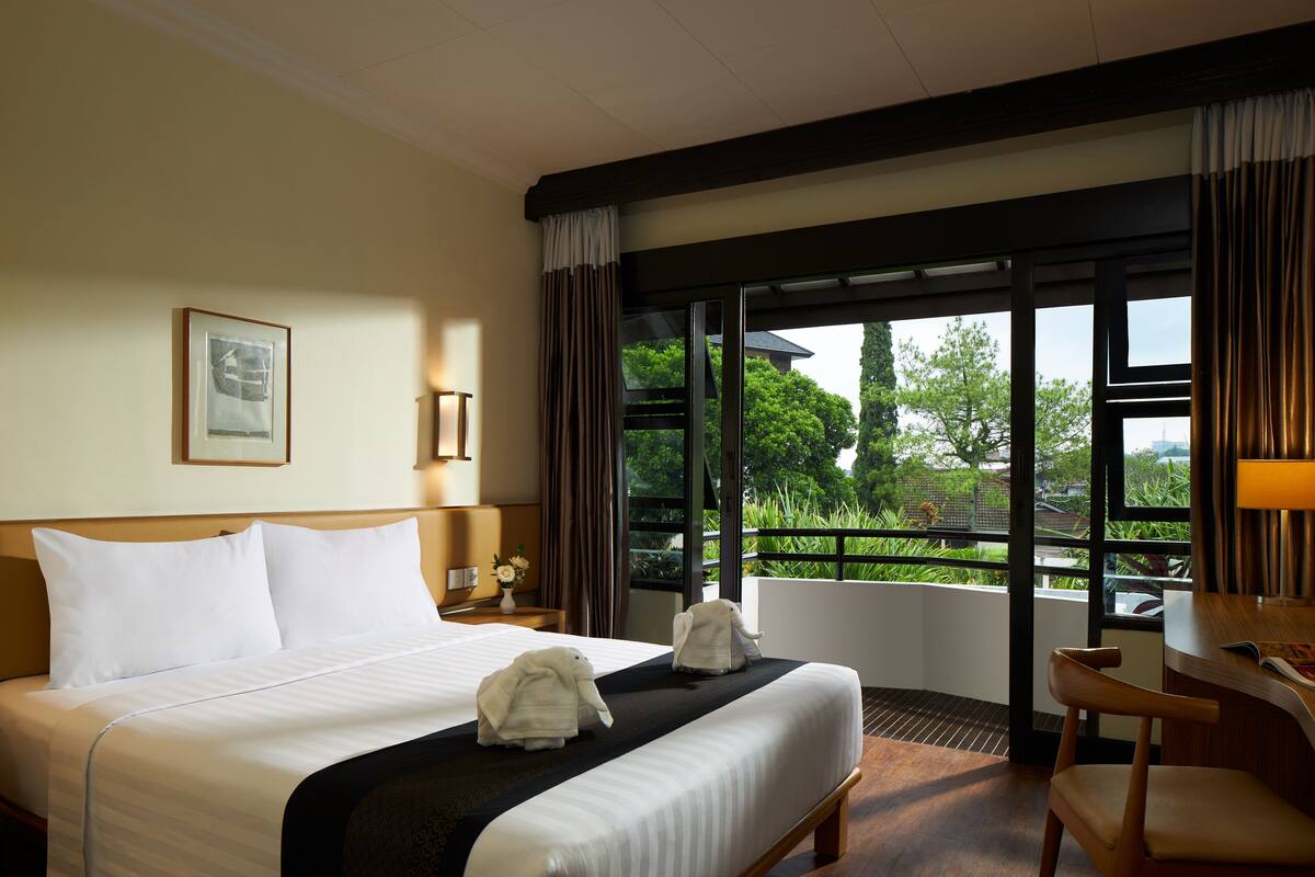 A hotel room with a large bed, two towels, a desk, and a view of greenery outside.