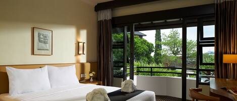 A hotel room with a large bed, two towels, a desk, and a view of greenery outside.