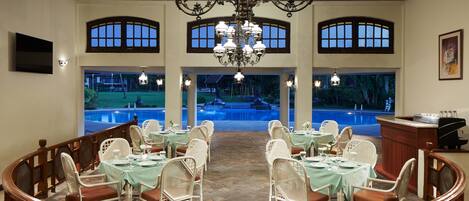 Uma sala de jantar com mesas preparadas para hóspedes, um candelabro e vista para uma piscina.