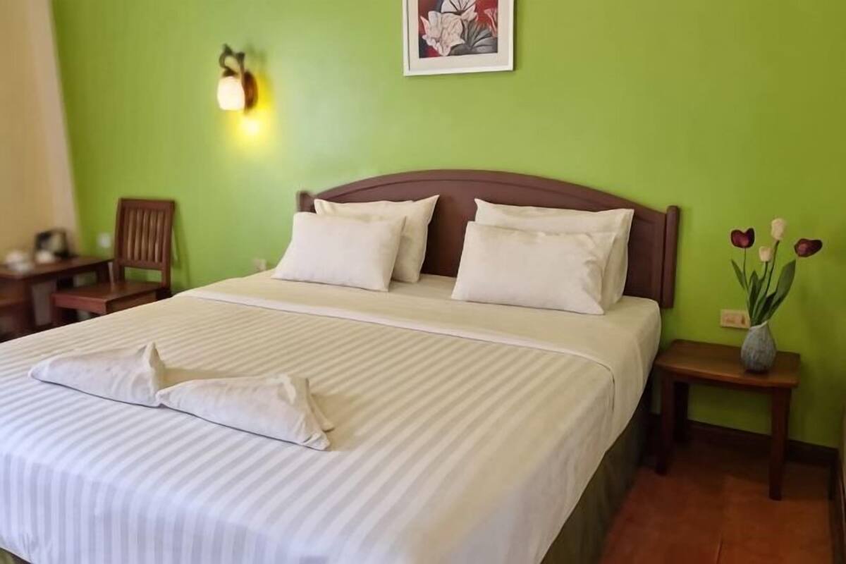 A neatly made bed with pillows, a bedside table with a vase of flowers, and a chair in a room with green walls.