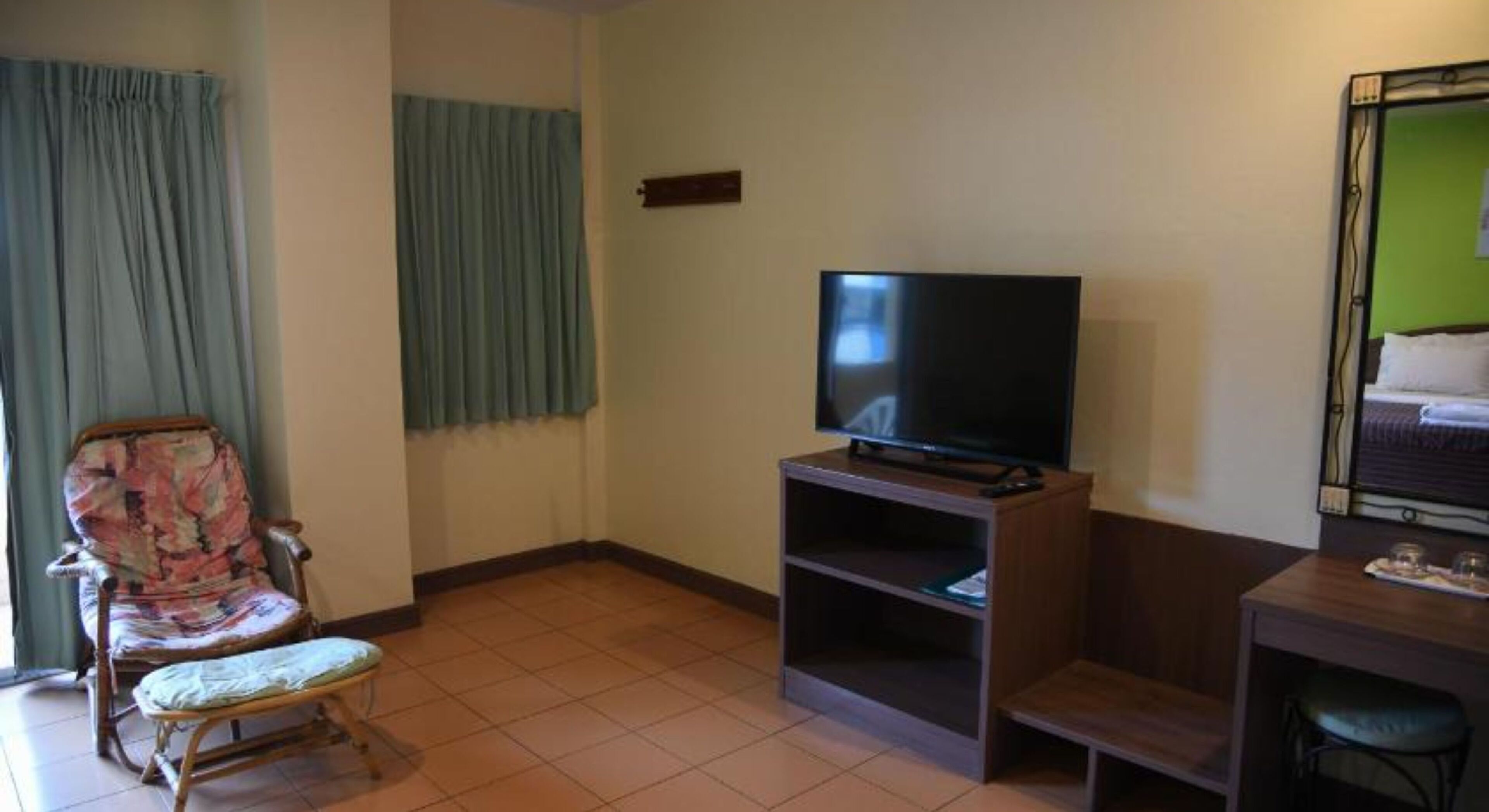 A room with a television on a wooden stand, a chair with a patterned cushion, and a bed with a green wall behind it.