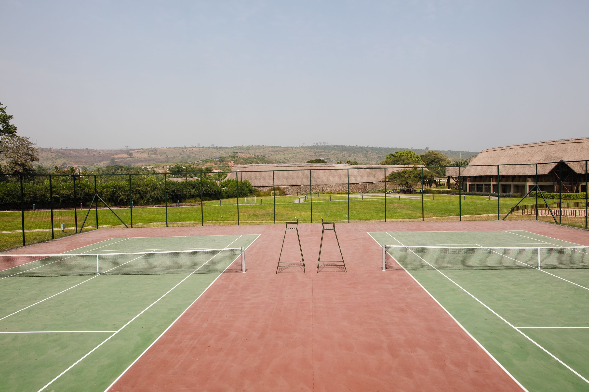 An outdoor tennis court with two courts, surrounded by a fence, and a building in the background.