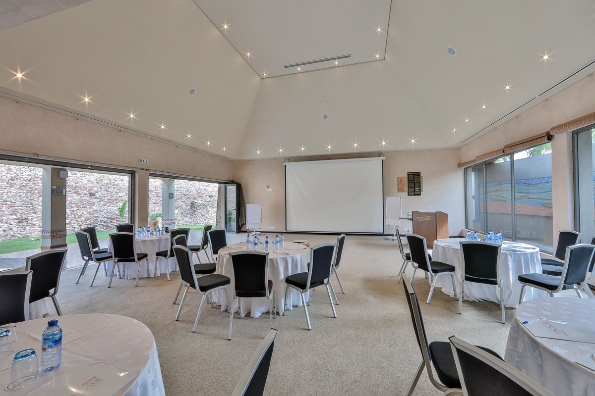 Un salón de conferencias con varias mesas y sillas, una pantalla de proyector y ventanas grandes que dan al exterior.