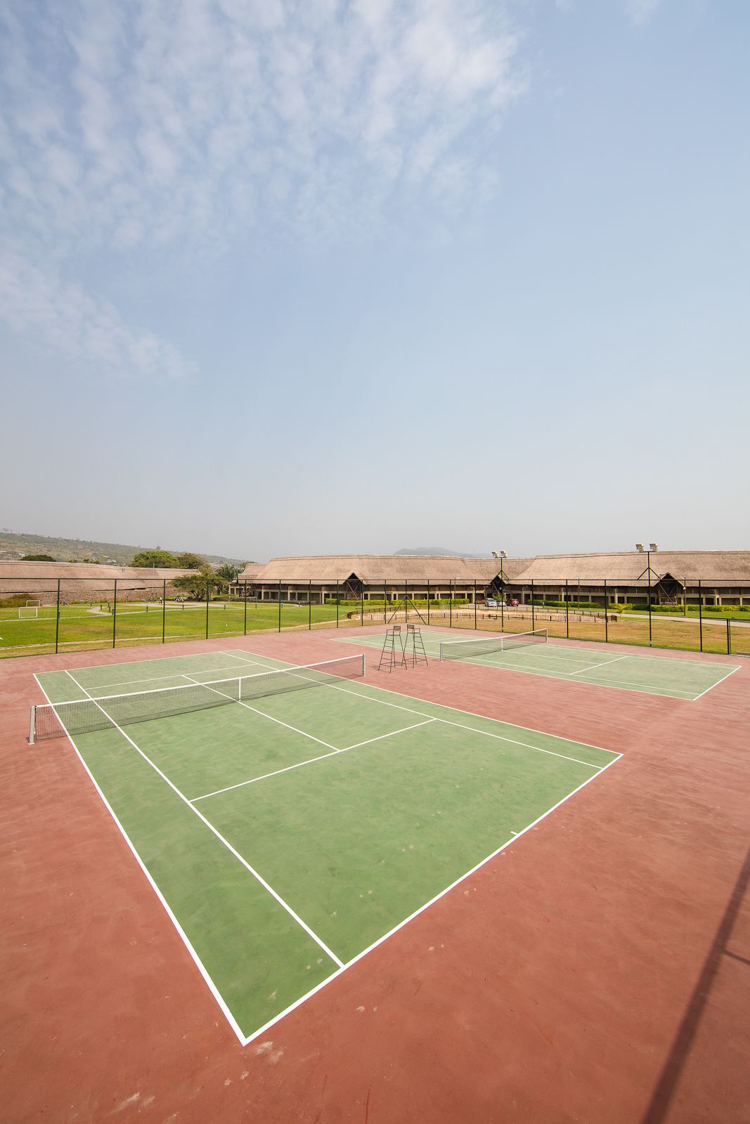 An outdoor tennis court with a net, surrounded by a fence, and a building in the background.