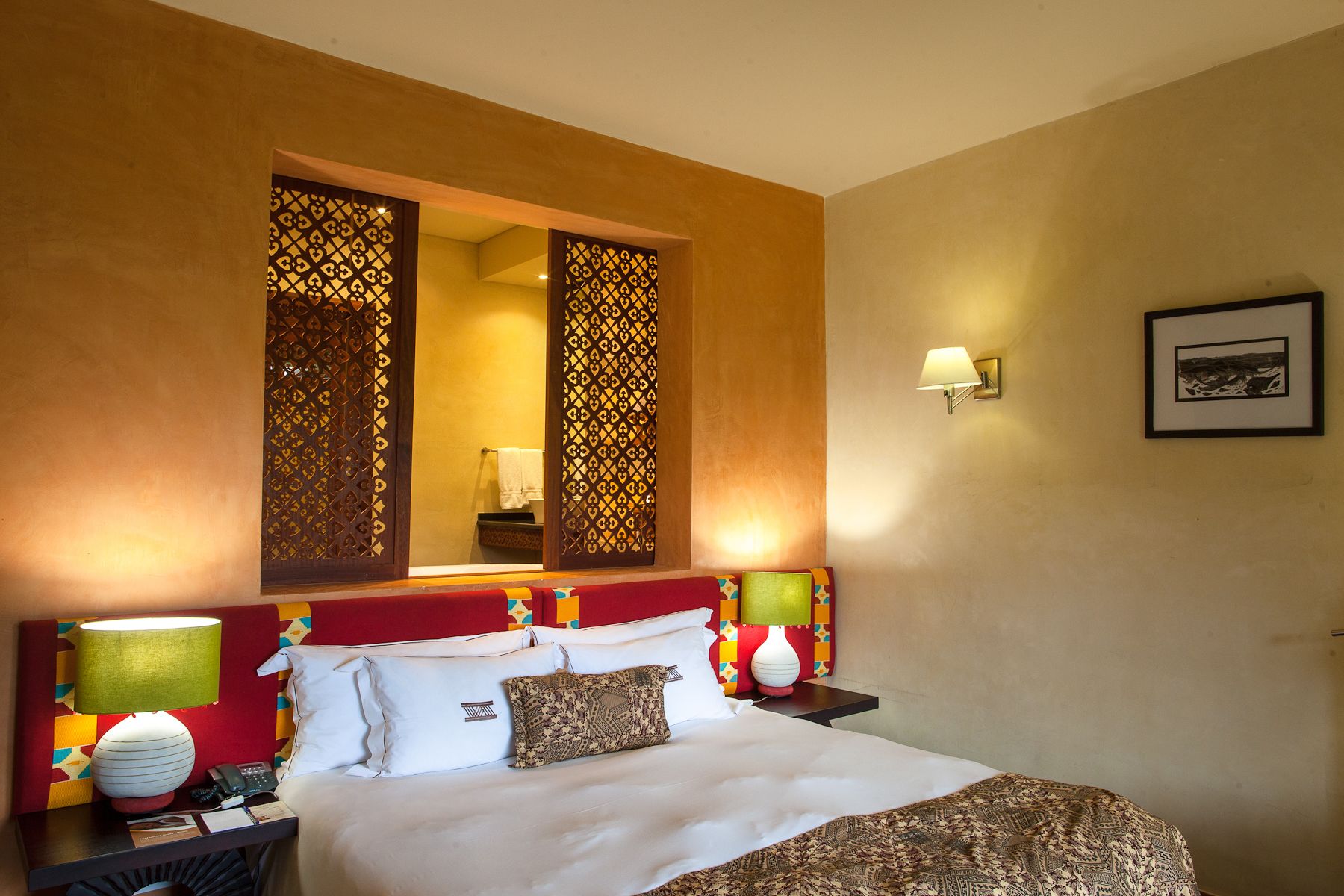 Quarto de hotel com cama, mesas de cabeceira, luminária, espelho e luminária fixada na parede.