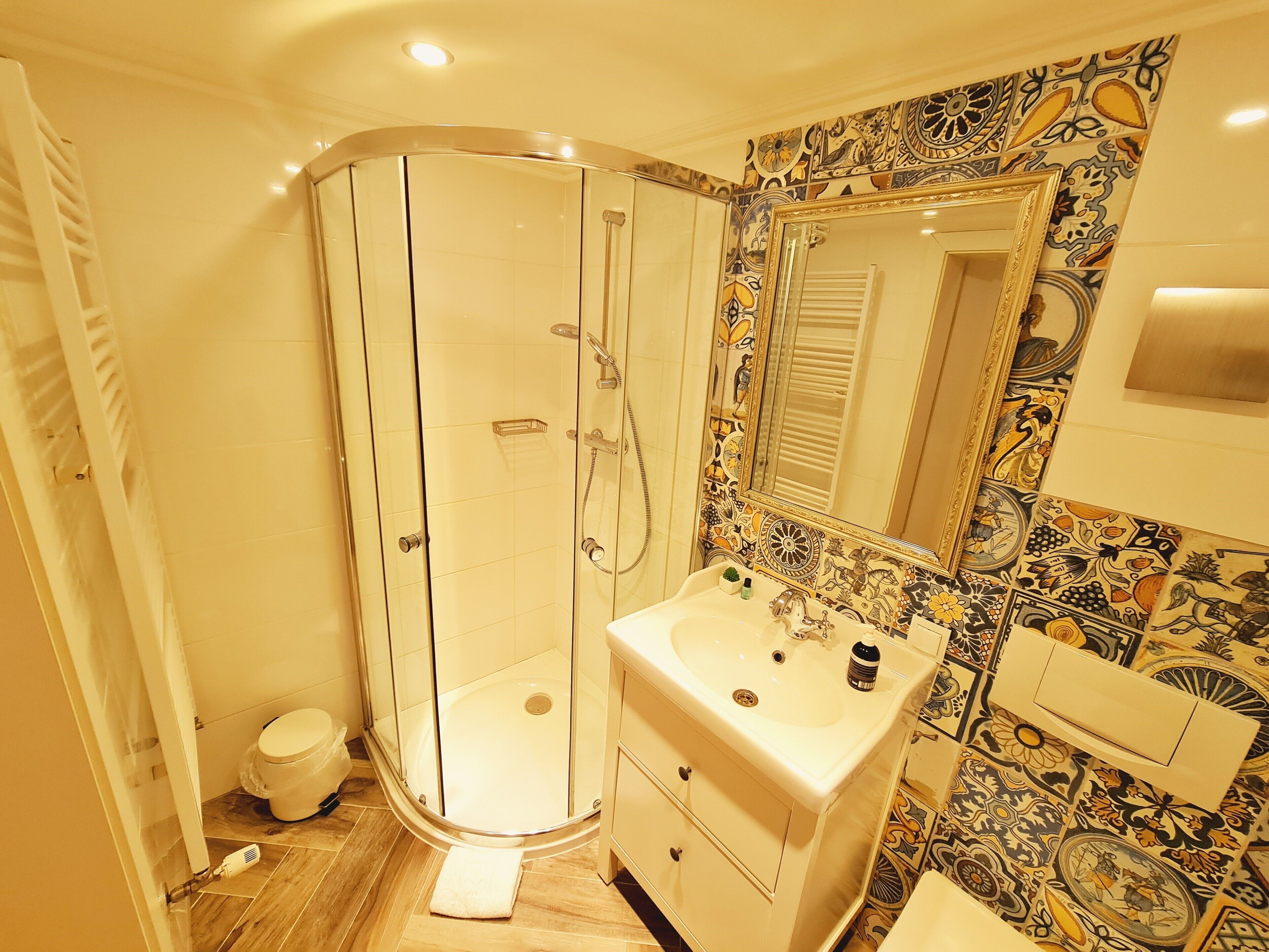 Een badkamer met een douchecabine van glas, een wit wastafelmeubel met een spiegel erboven en een betegelde muur met een decoratief patroon.