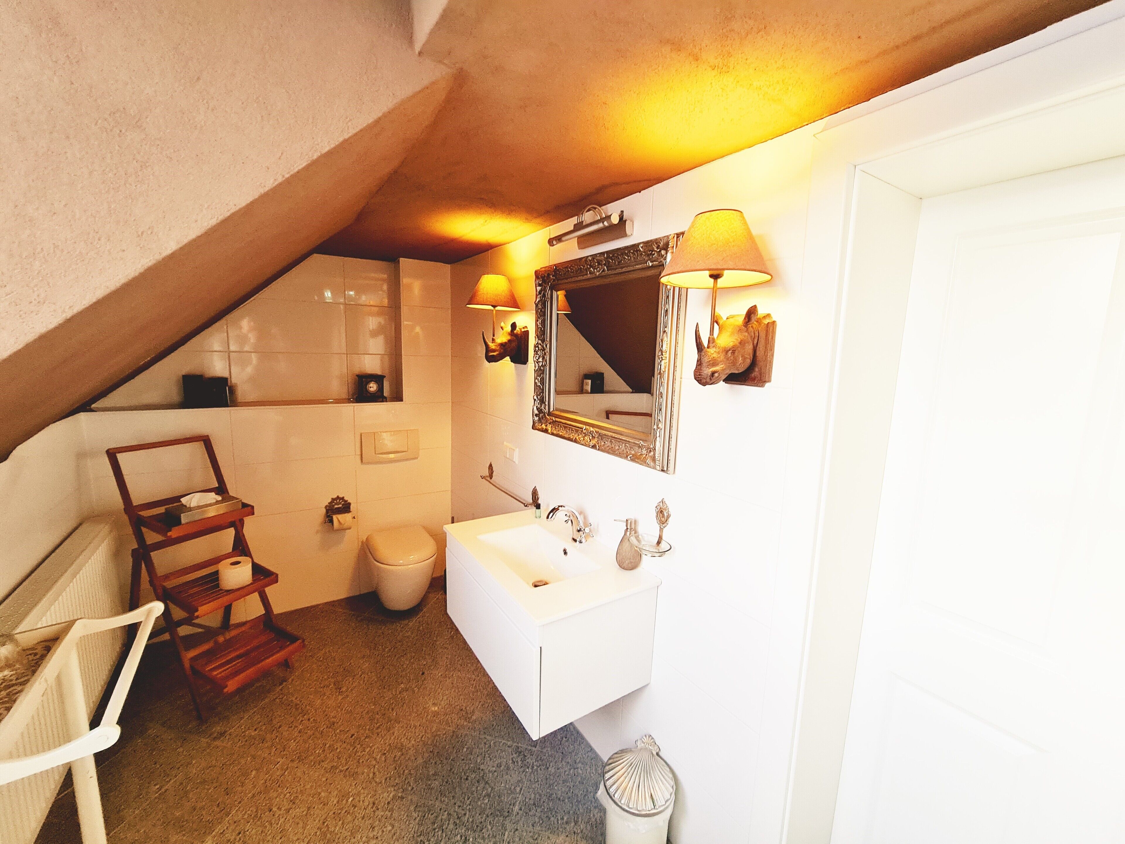 A bathroom with a white sink, mirror, and toilet. There is a wooden chair and a small shelf with toiletries.