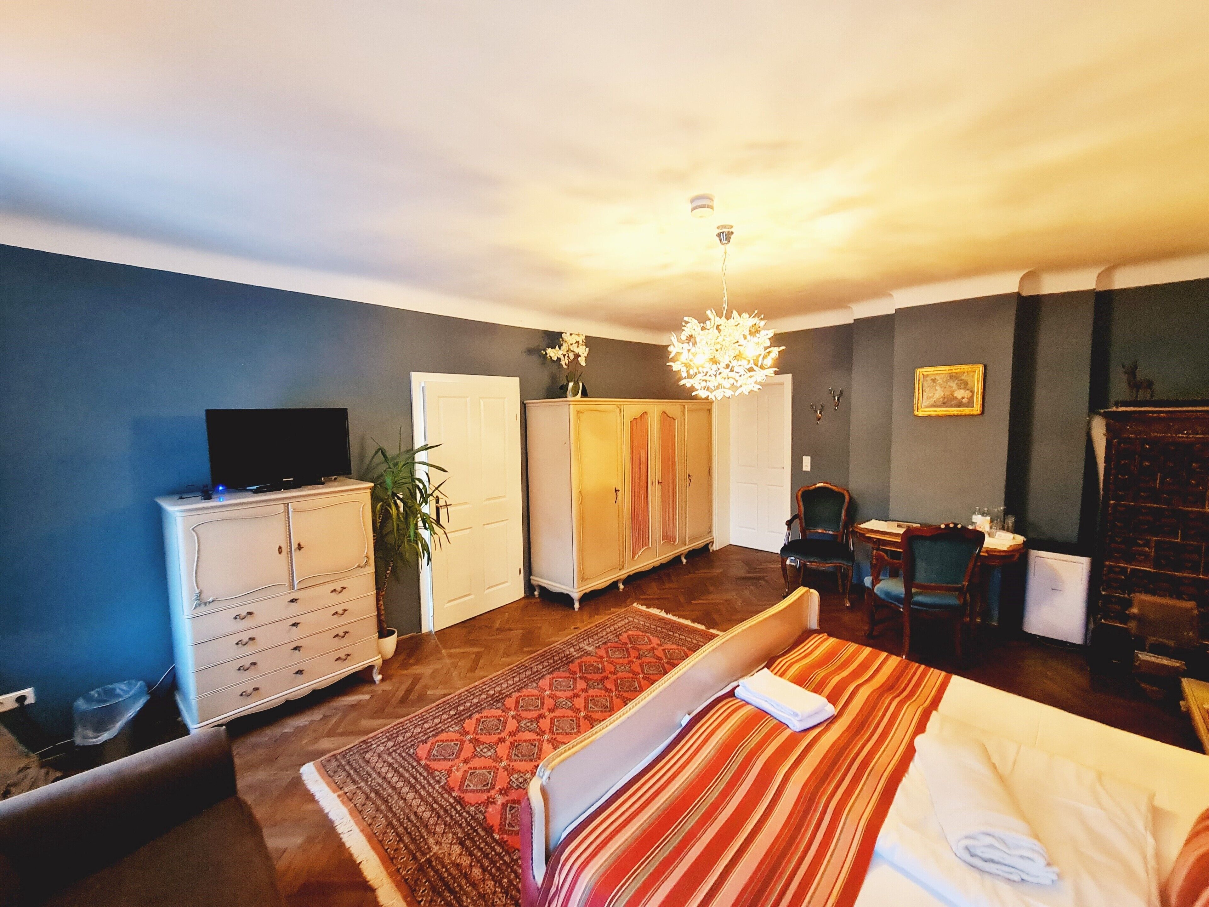A hotel room with a bed, a television on a dresser, a wardrobe, a desk with a chair, and a chandelier.