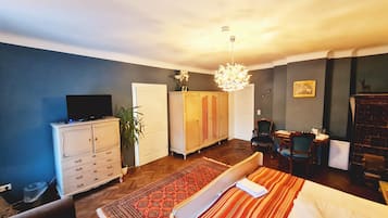 A hotel room with a bed, a television on a dresser, a wardrobe, a desk with a chair, and a chandelier.