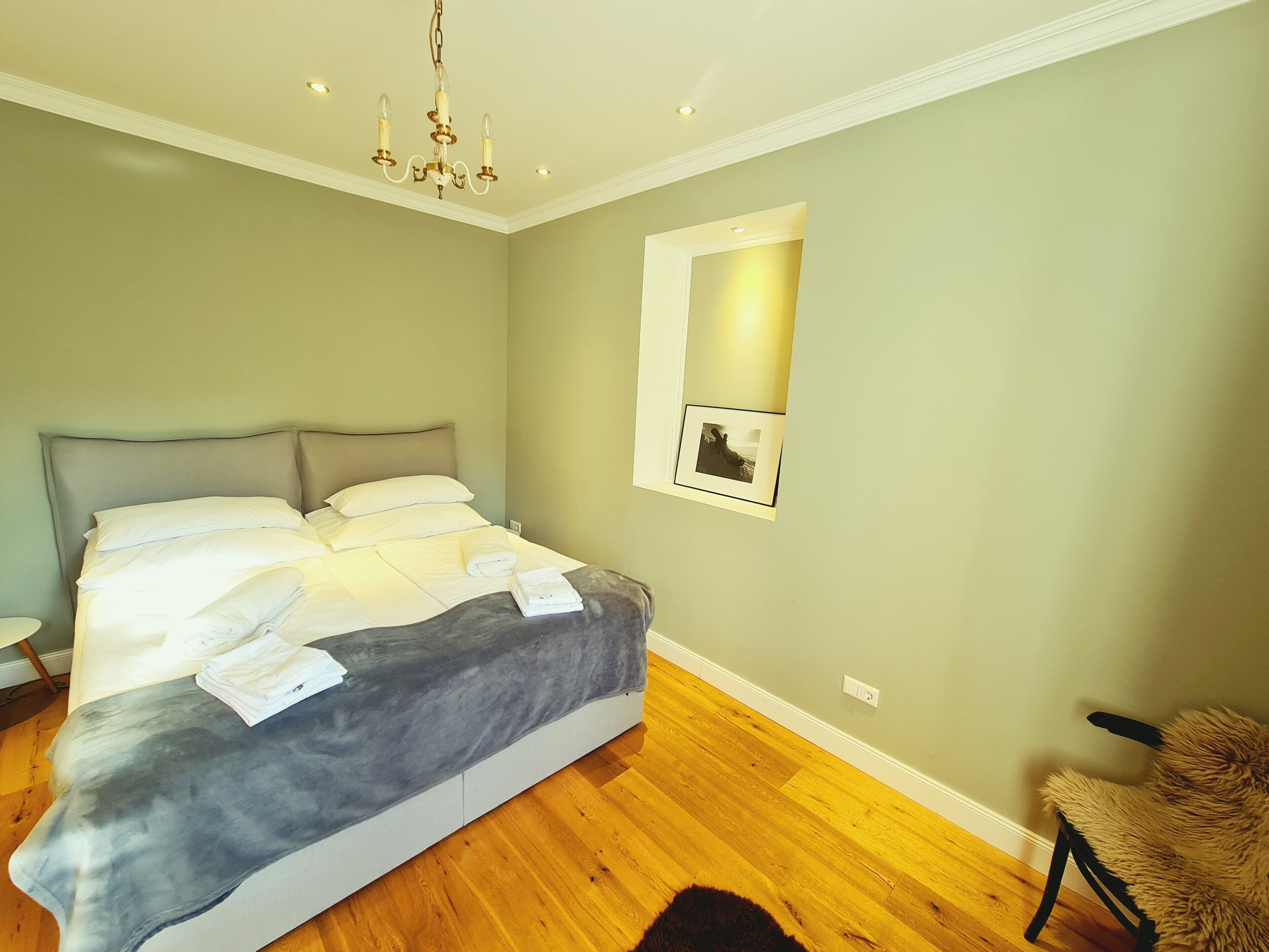 Een net opgemaakt bed met witte lakens en een grijze deken, een houten vloer, een kroonluchter en een ingelijst schilderij aan de muur.