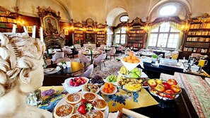 A library with ornate woodwork, bookshelves, and a dining area with fruit and pastries.