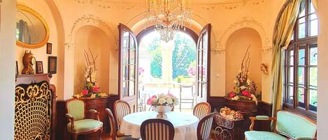 An ornate room with a chandelier, a dining table with chairs, and a large window with a view of greenery.