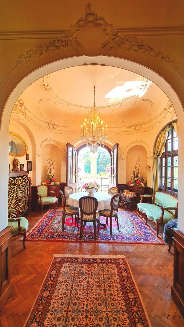 An ornate room with a chandelier, a dining table with chairs, and a large window with a view of greenery.