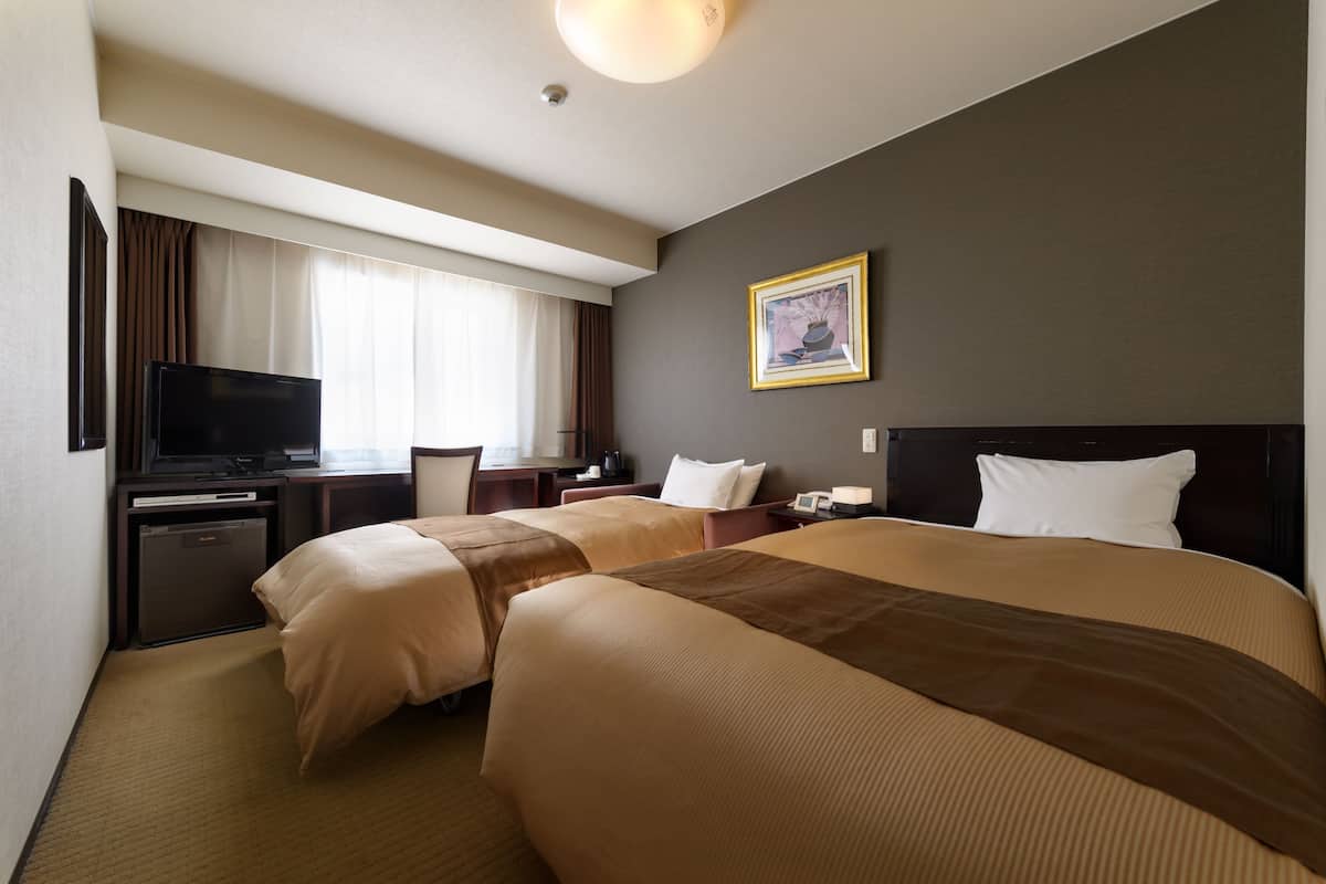 Una habitación de hotel con dos camas, un escritorio con televisor, un refrigerador pequeño y un cuadro en la pared.