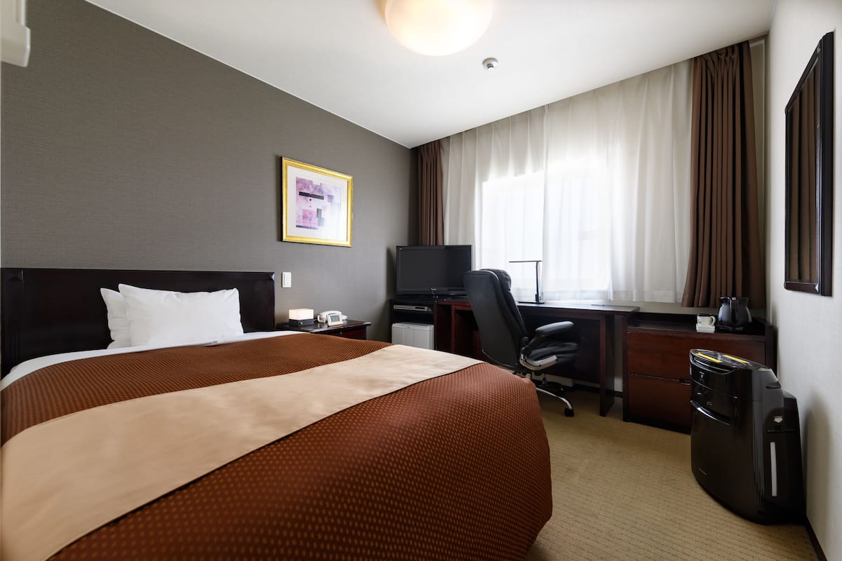Habitación de hotel con una cama grande, un escritorio con computadora, una silla, una cafetera y una maleta.