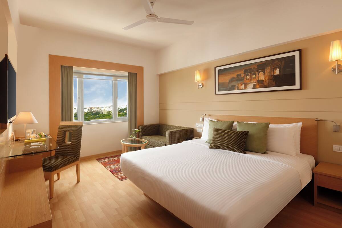Ein Hotelzimmer mit einem großen Bett, einem Schreibtisch mit Lampe, einem Sessel, einem kleinen Tisch und einem Fenster mit Stadtblick.