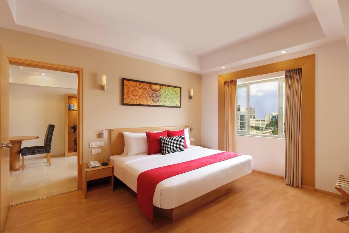 Ein Hotelzimmer mit einem großen Bett, einem Schreibtisch, einem Sessel, einem Fenster mit Vorhängen, einem Bild an der Wand und einem Fernseher.