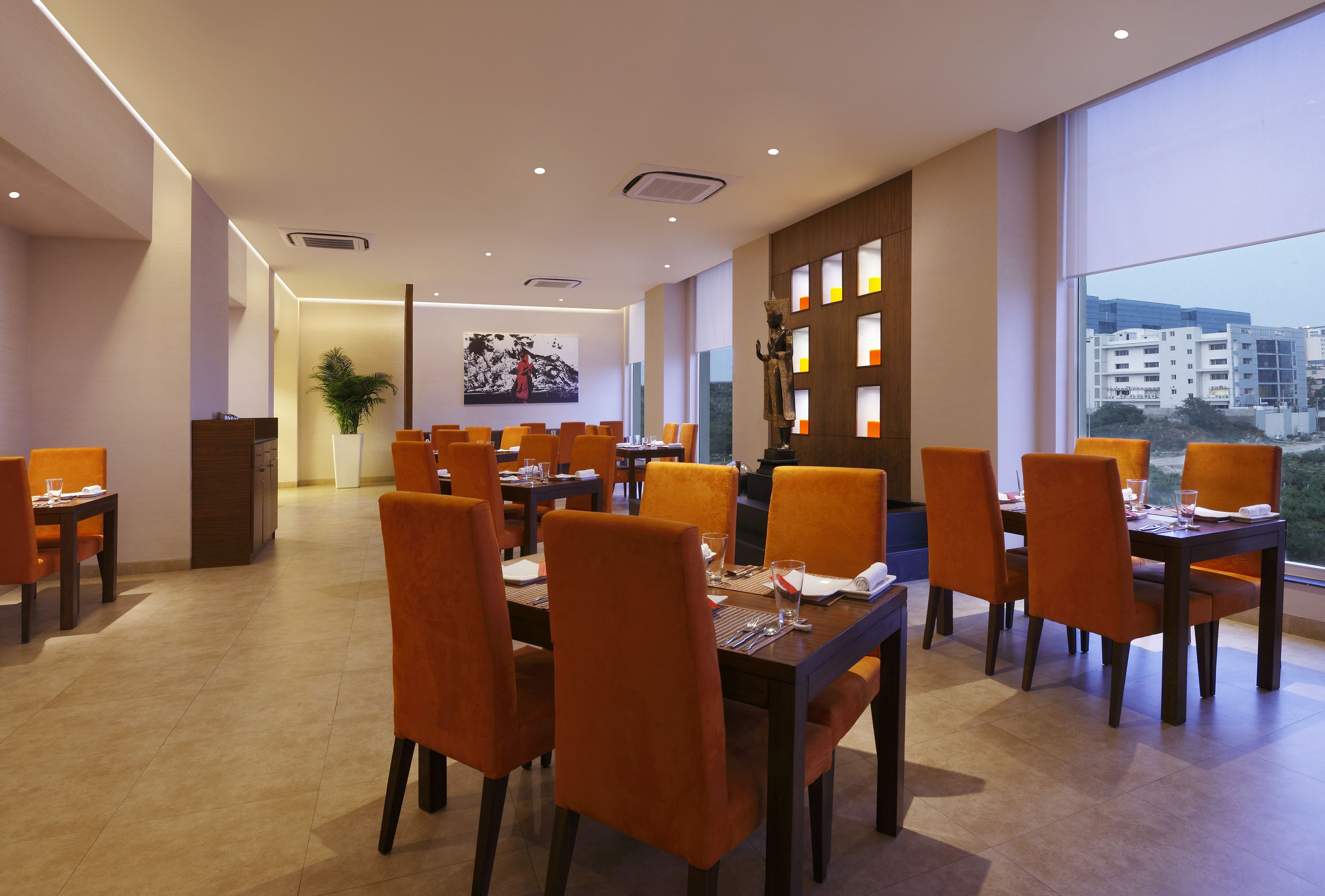 Un espace de restauration moderne avec des chaises et des tables orange, une grande fenêtre donnant sur la ville et un tableau accroché au mur.