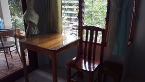 A dining area with a wooden table and chairs, a window with curtains, and a view of greenery outside. - Pattara House (Koh Samui)