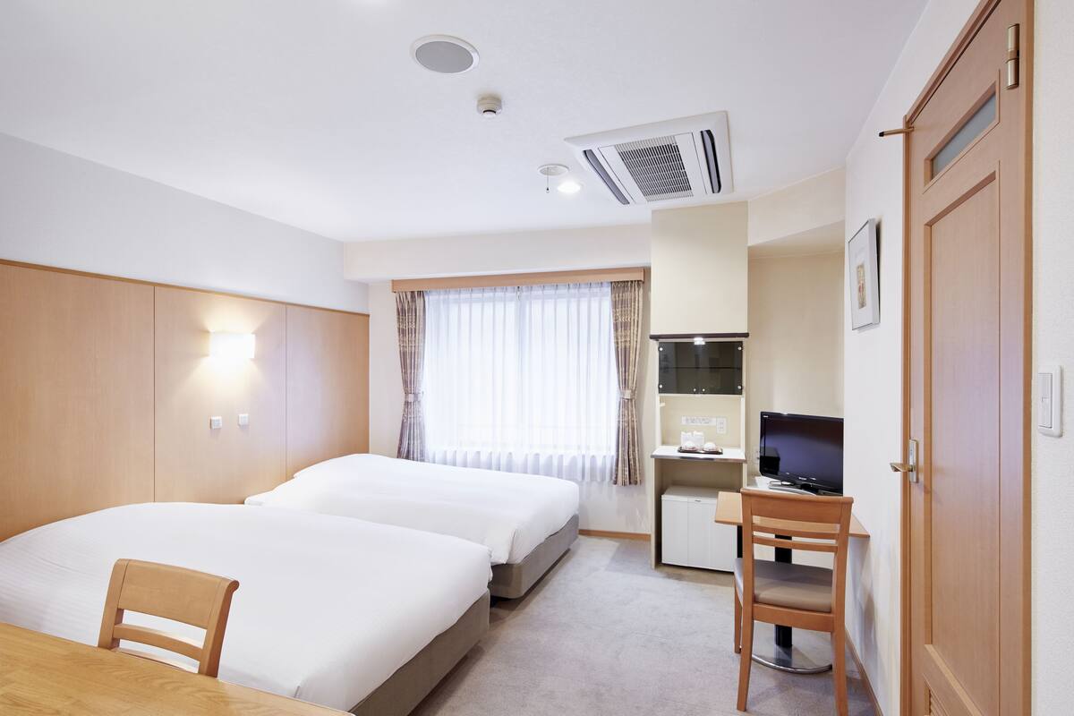 Une chambre d’hôtel avec deux lits, un bureau avec un ordinateur, une petite kitchenette et une fenêtre avec des rideaux.