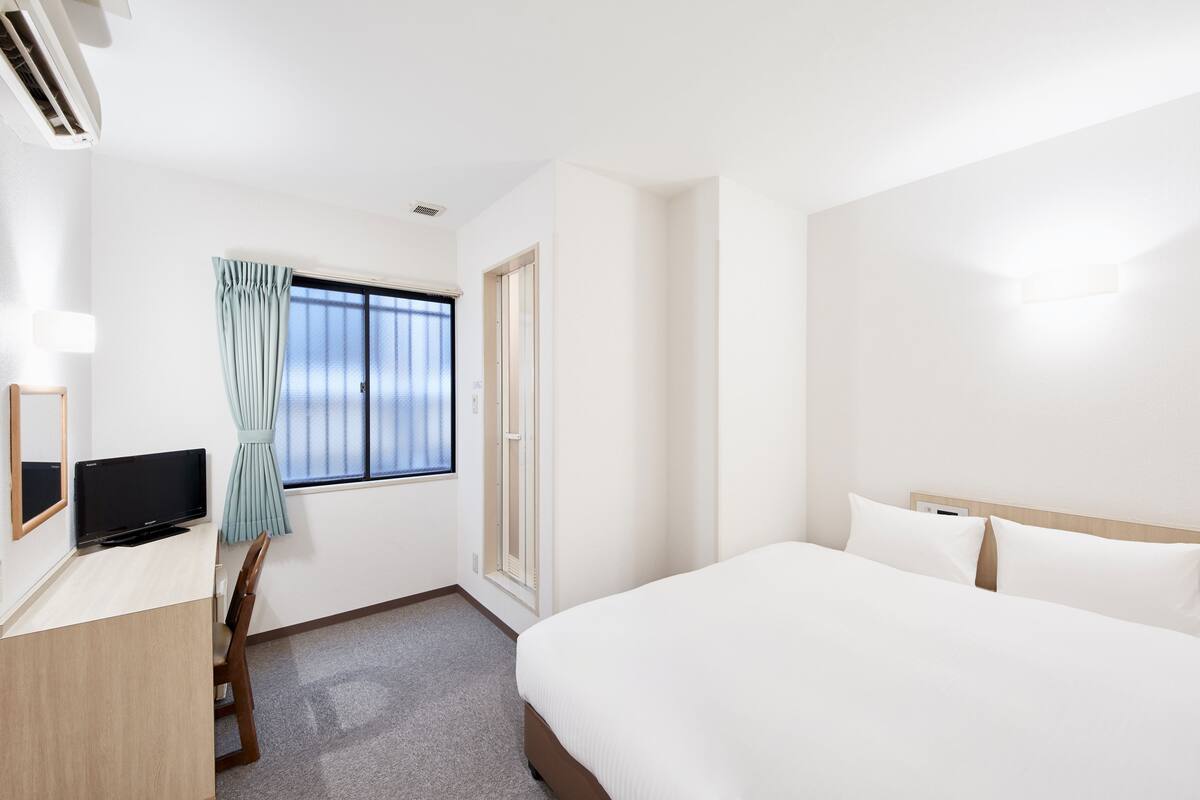Une chambre d’hôtel avec un lit, un bureau avec une télévision, une fenêtre avec des rideaux et une porte.
