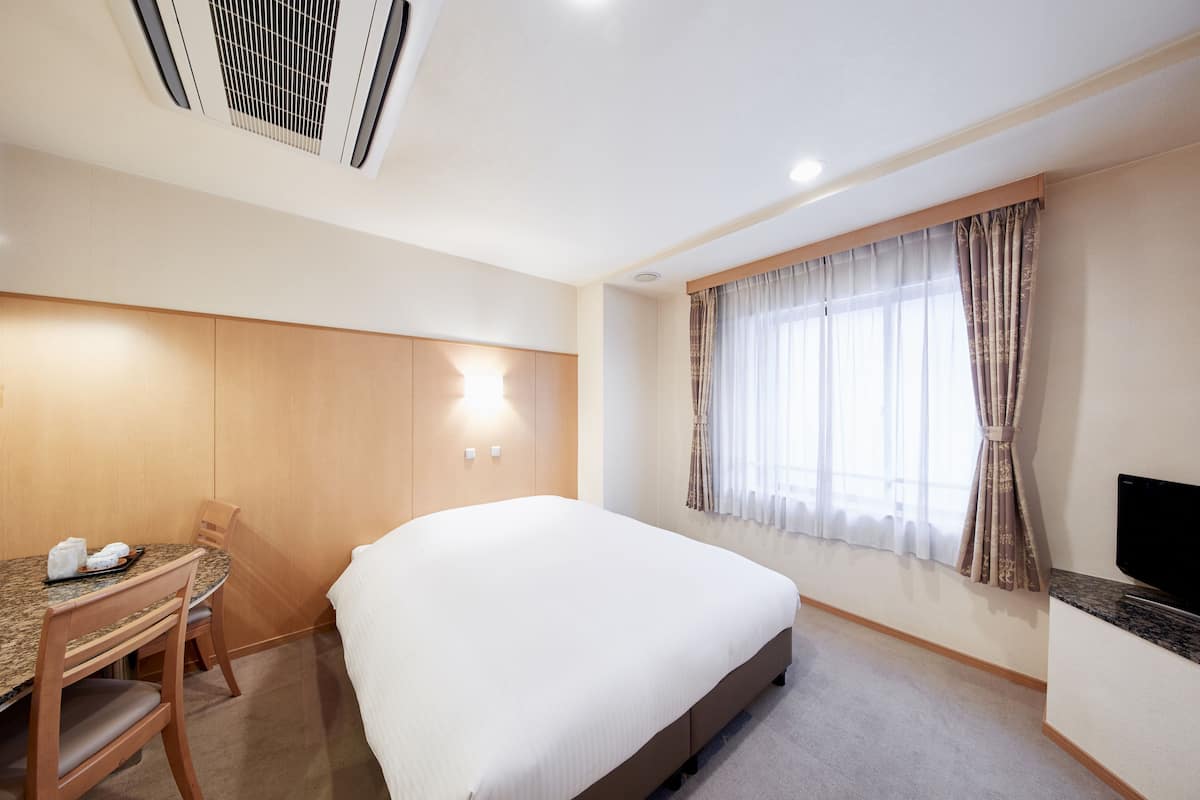 Une chambre d’hôtel comprenant un lit, un bureau avec une chaise, une télévision et une fenêtre avec des rideaux.