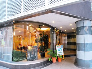 Entrada de hotel con mostrador de recepción, plantas en macetas y un cartel.