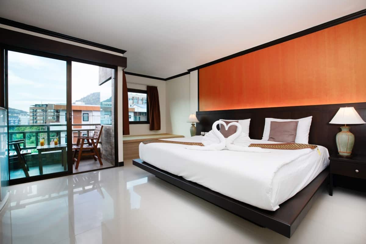 A modern hotel room with a large bed, a balcony with a view, and a cityscape visible through the windows.