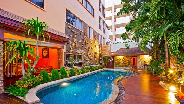 Una piscina con un muro de piedra y plantas, rodeada por una terraza de madera y un edificio con múltiples ventanas.