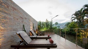 Un balcón con dos sillones de descanso, baranda de vidrio y vistas a zonas verdes y edificios al lejano.