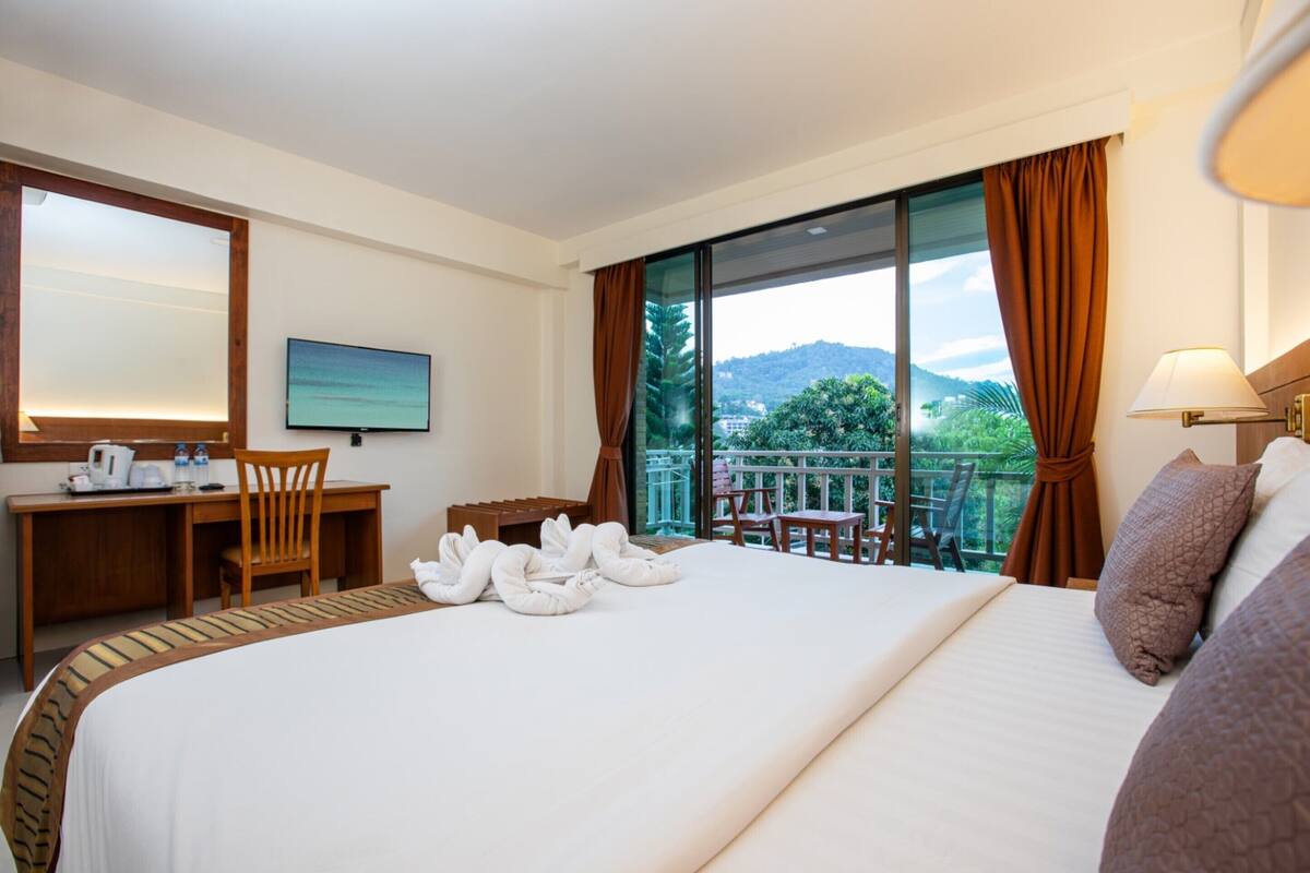 A hotel room with a large bed, a desk with a chair, a television, and a balcony with a view of greenery and mountains.