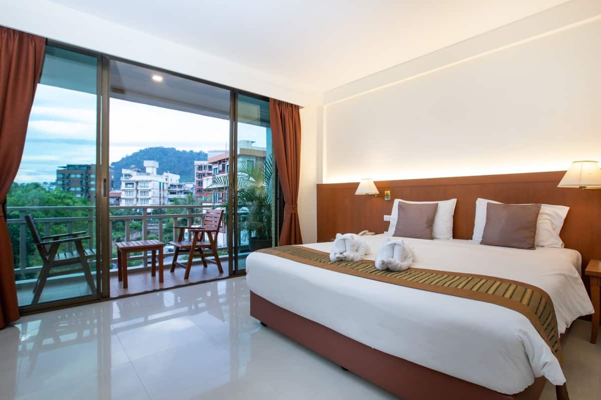 A hotel room with a large bed, a balcony with chairs, and a view of buildings and hills.