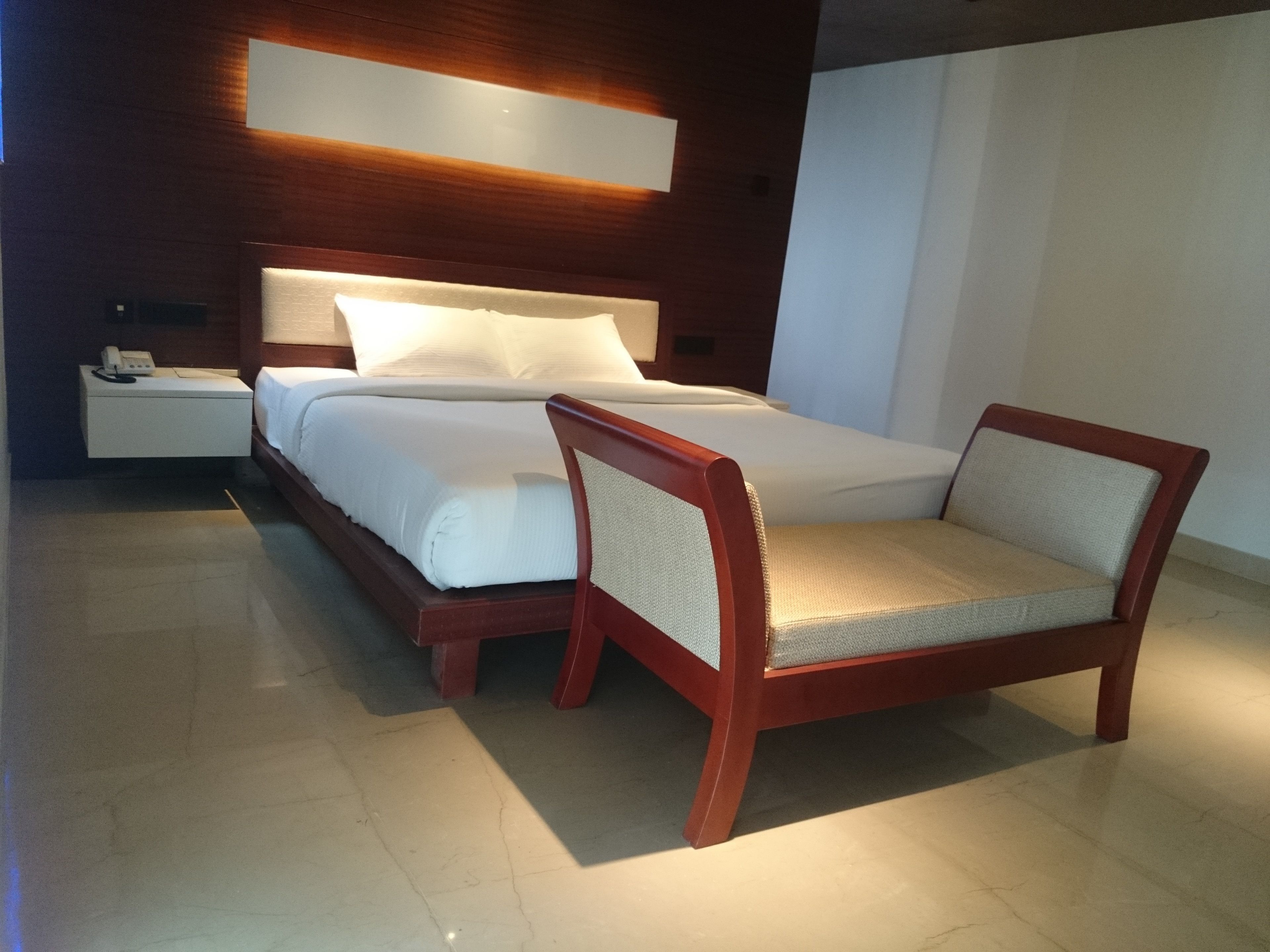 A modern hotel room with a neatly made bed, a wooden nightstand with a phone, and a wooden chair.