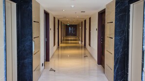Corredor longo e bem iluminado, com piso de mármore e portas em ambos os lados, que leva a uma saída no final.
