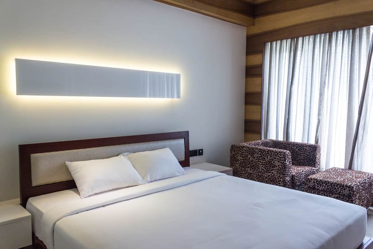 Habitación de hotel con una cama grande, cabecera, mesita de noche, una silla y ventana con cortinas.