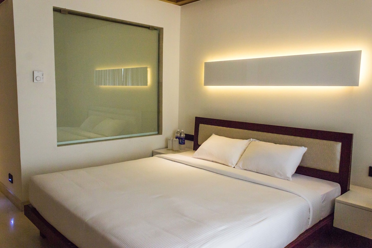 Una habitación de hotel con una cama grande, una mesita de noche, una lámpara de pared y una ventana con persianas.