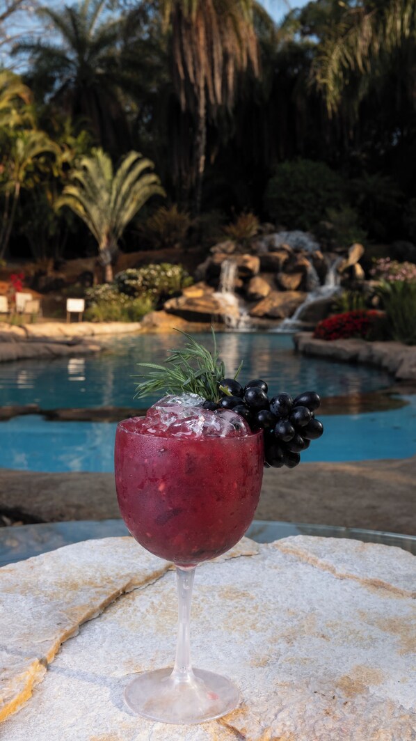 A glass of red beverage with a garnish, placed on a stone surface near a pool with a waterfall and palm trees in the background. - Ville Real Hotel (Ouro Preto)
