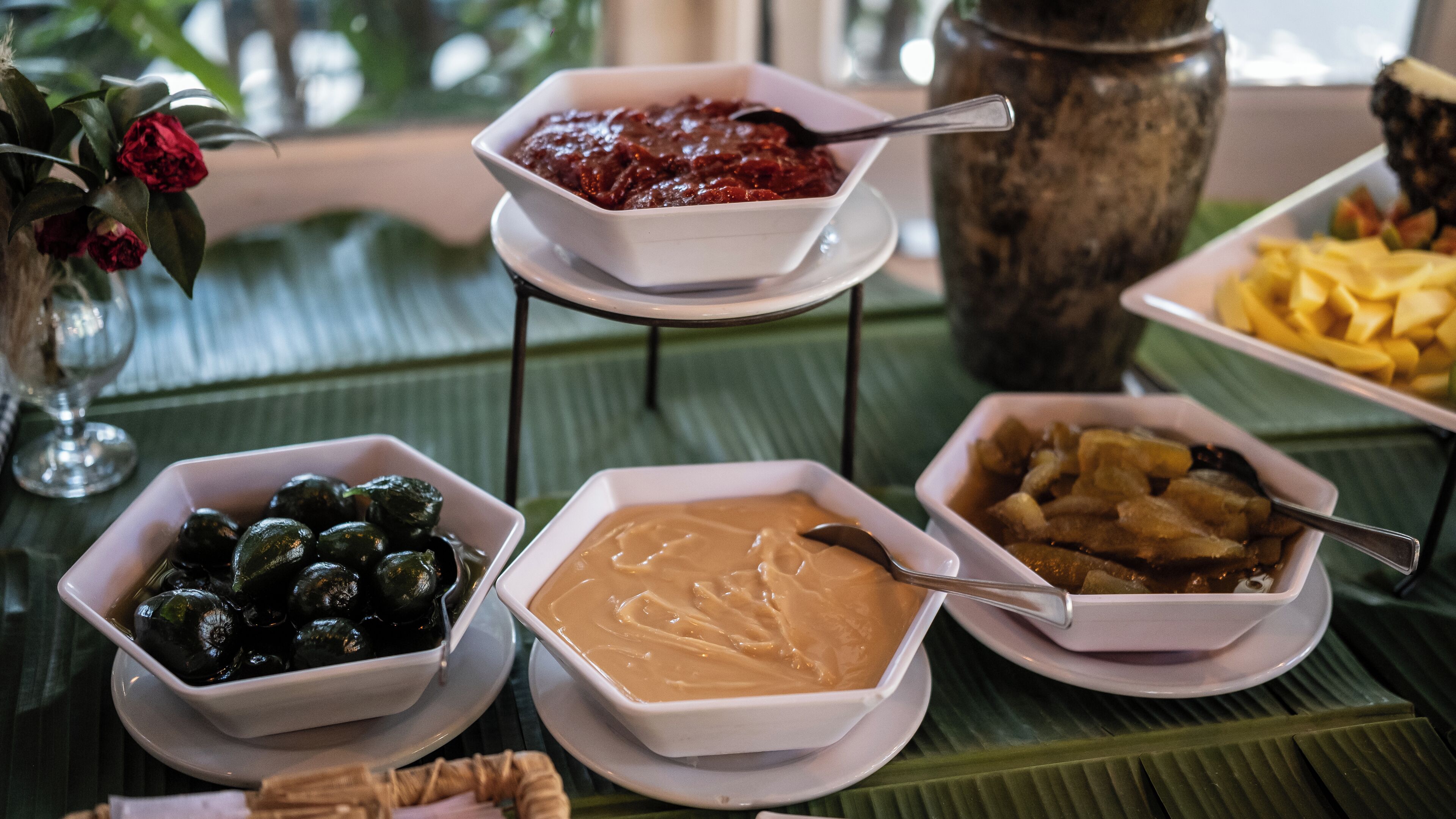 Una mesa con varios platos, incluyendo un bol de salsa oscura, un bol de salsa cremosa, un bol de aceitunas verdes y un plato de fruta amarilla.
