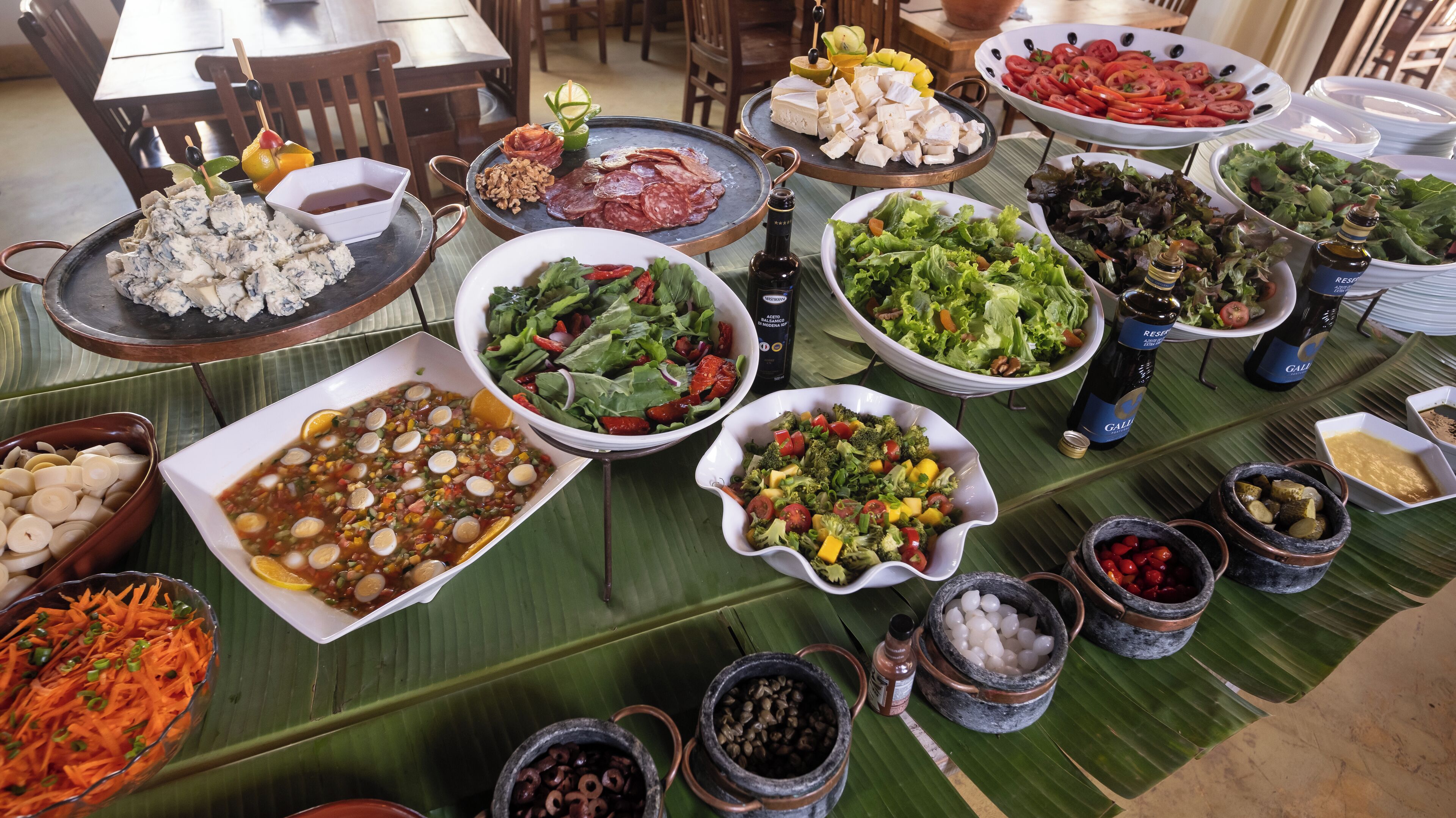 Uma mesa de buffet com diversos pratos, incluindo saladas, carnes e legumes, dispostos sobre folhas de bananeira.