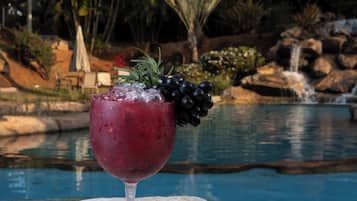 A glass of red drink with grapes on a stone surface by a pool with palm trees and a waterfall in the background.