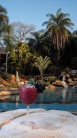 Un vaso de bebida roja con uvas sobre una superficie de piedra junto a una piscina con palmeras y una cascada al fondo.
