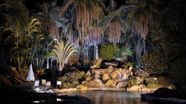 Vista nocturna de un jardín ajardinado con cascada, palmeras y zona de piscina.