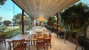 Un área de comedor al aire libre con mesas y sillas de madera, rodeada de plantas y vista del paisaje.