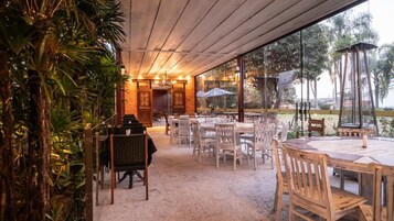 Un área de comedor al aire libre con varias mesas y sillas, rodeada por paredes de vidrio y plantas tropicales.