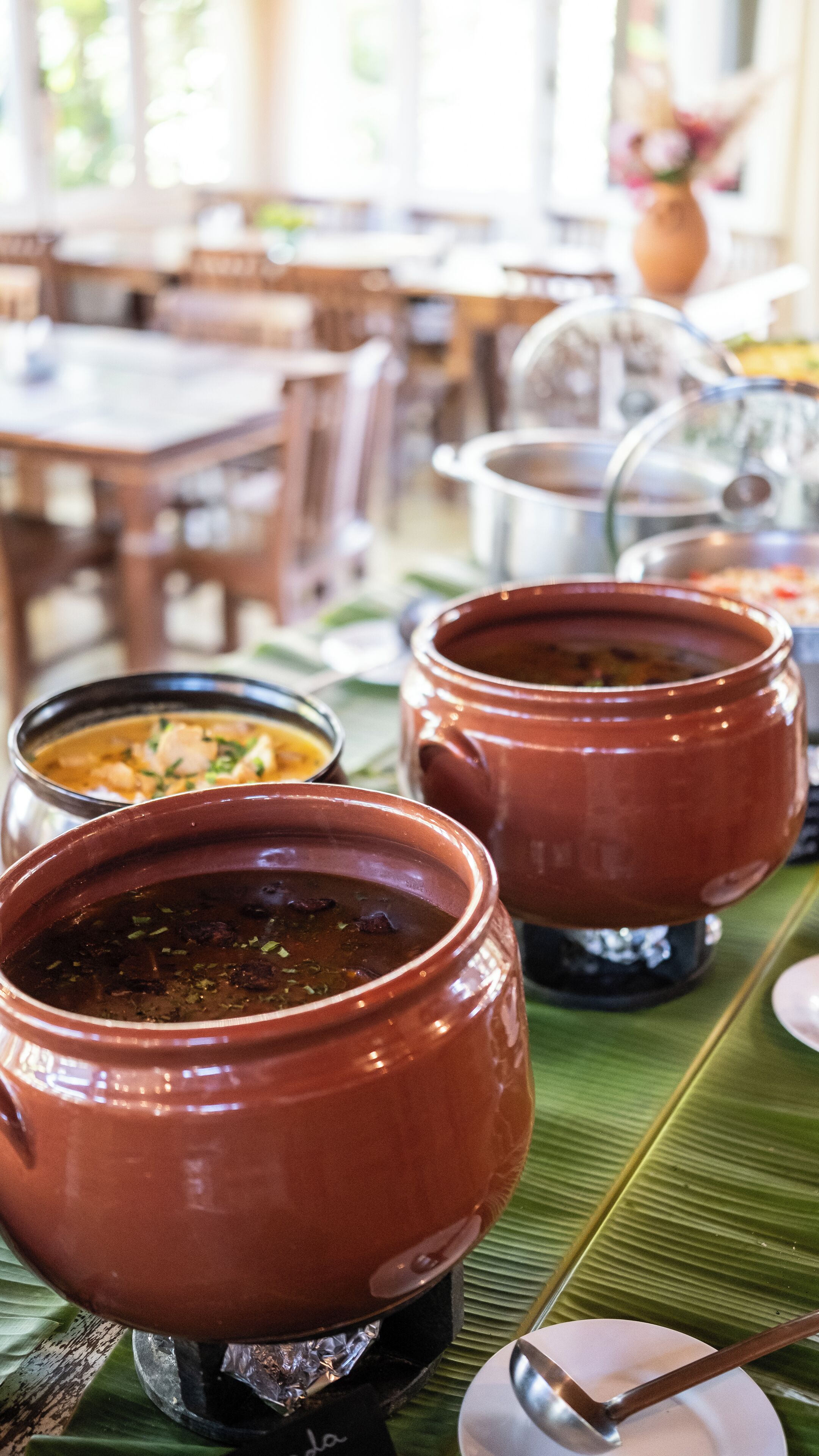 A table with two large clay pots of soup, a smaller bowl of soup, and a spoon on a banana leaf.