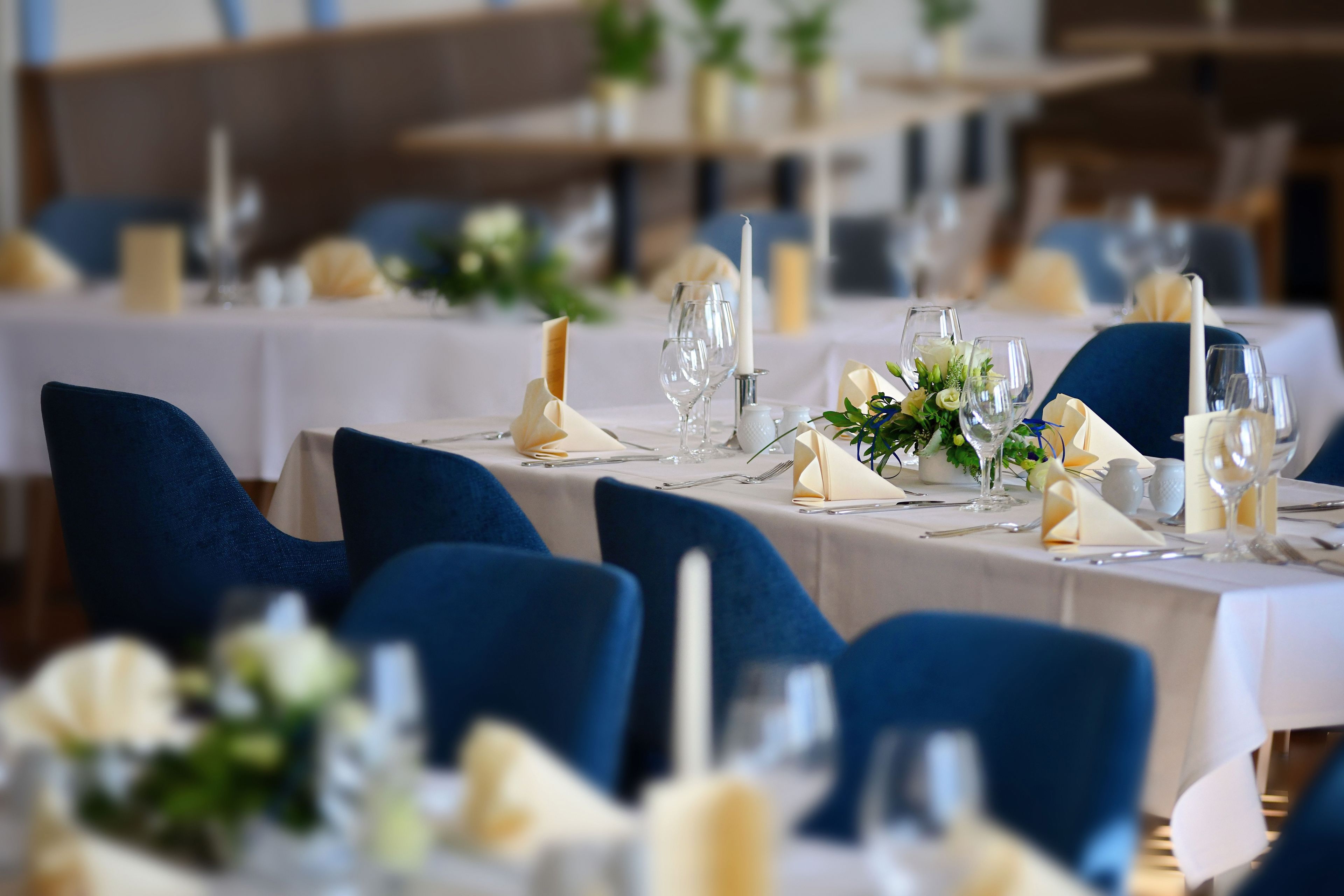 Een eetruimte met gedekte tafels voor een formeel evenement, met blauwe stoelen, witte tafelkleden en bloemstukken als decoratie.