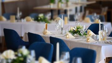 Een eetruimte met gedekte tafels voor een formeel evenement, met blauwe stoelen, witte tafelkleden en bloemstukken als decoratie.