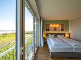 A bedroom with a large window, a bed, a bedside table, and a view of the beach.
