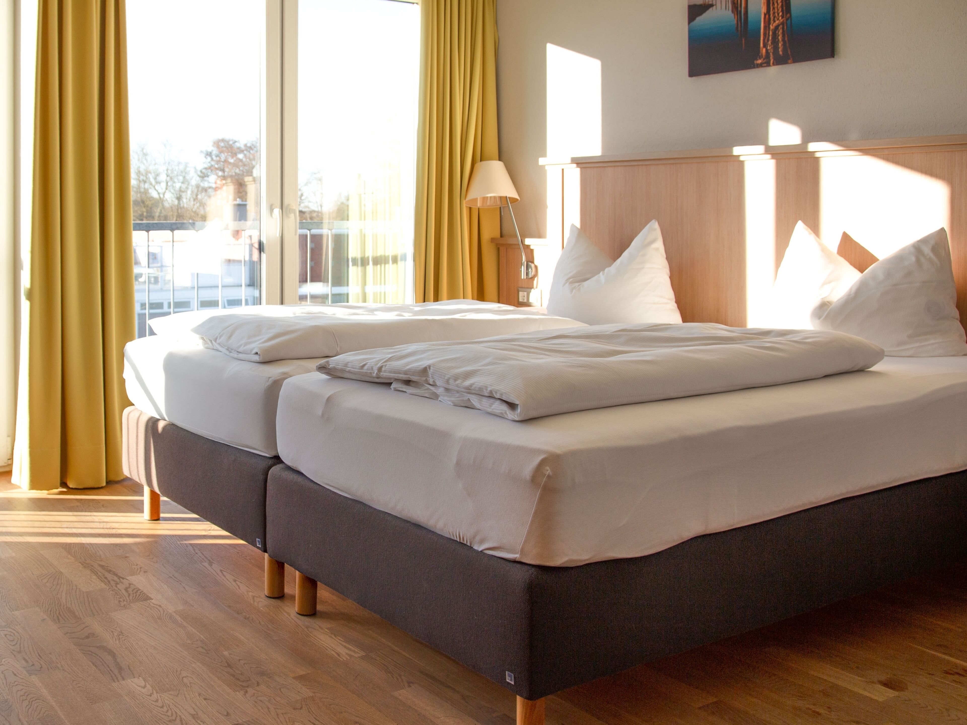 Ein Doppelbett mit weißer Bettwäsche, ein Holz-Kopfteil und ein Ausblick ins Freie durch große Fenster.
